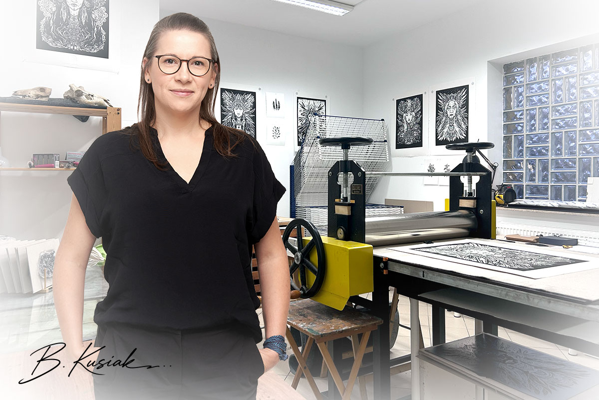 Barbara Kusiak w swojej pracowni graficznej, za nią prasa drukarska i odbitki linorytów na ścianie.
